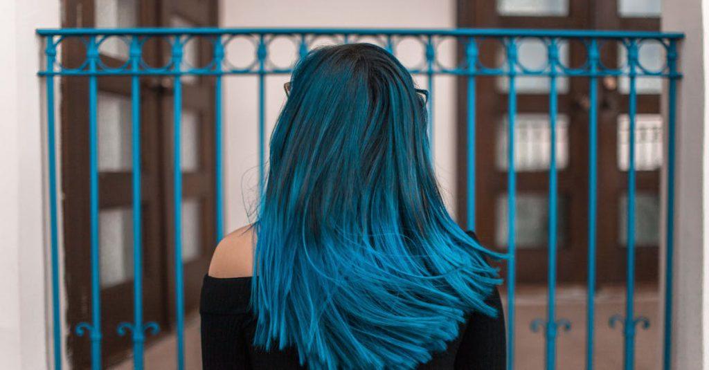 A young woman with vibrant blue hair stands against a stylish urban backdrop.